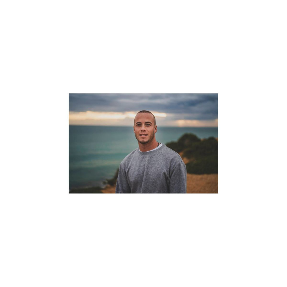 Ein Mann mit kurzen Haaren lächelt in die Kamera. Er trägt einen grauen Pullover, mit Meerblick.