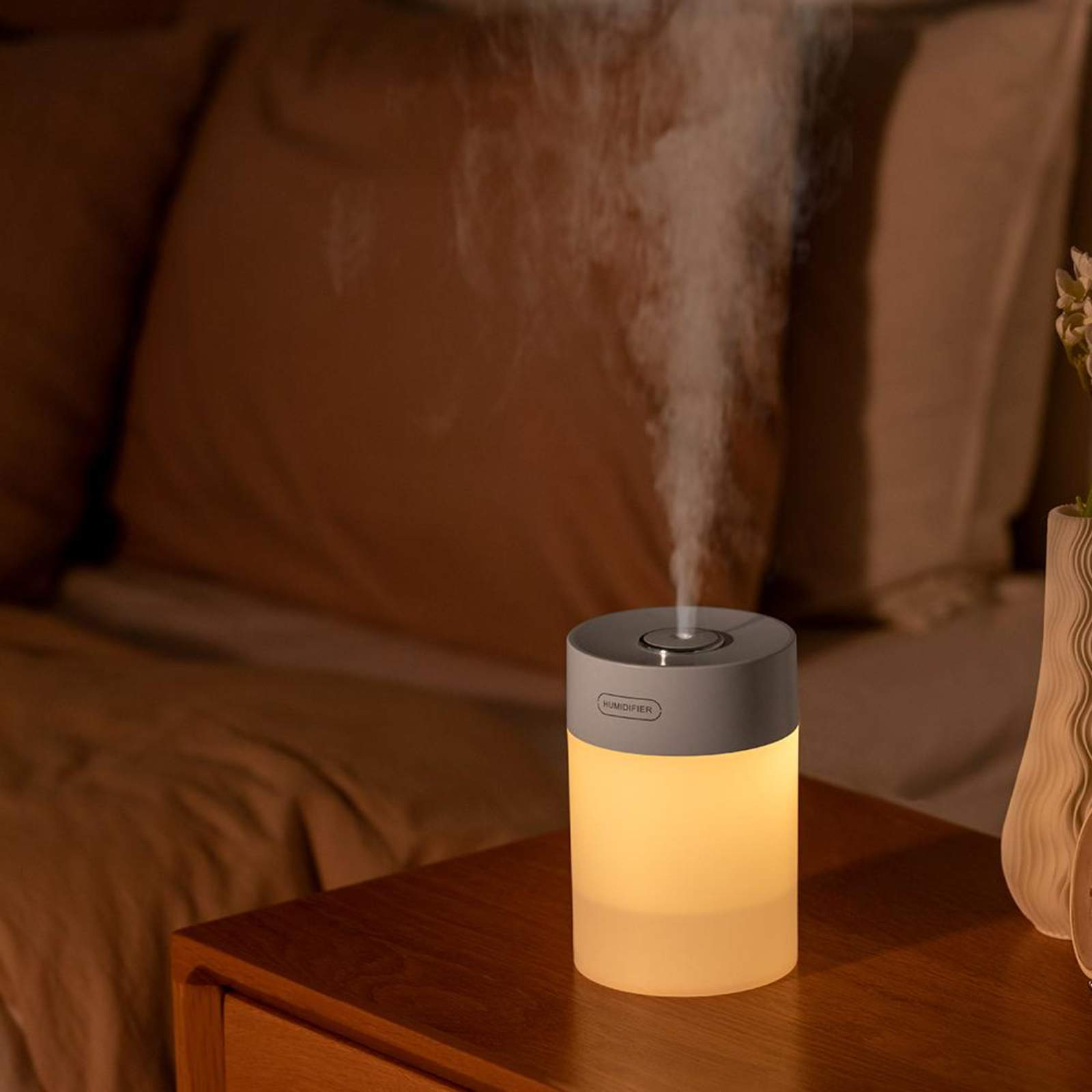 Ein kleiner, beleuchteter Luftbefeuchter auf einem Nachttisch, mit aufsteigendem Nebel. Schlafzimmer-Setting, sanfte Beleuchtung.