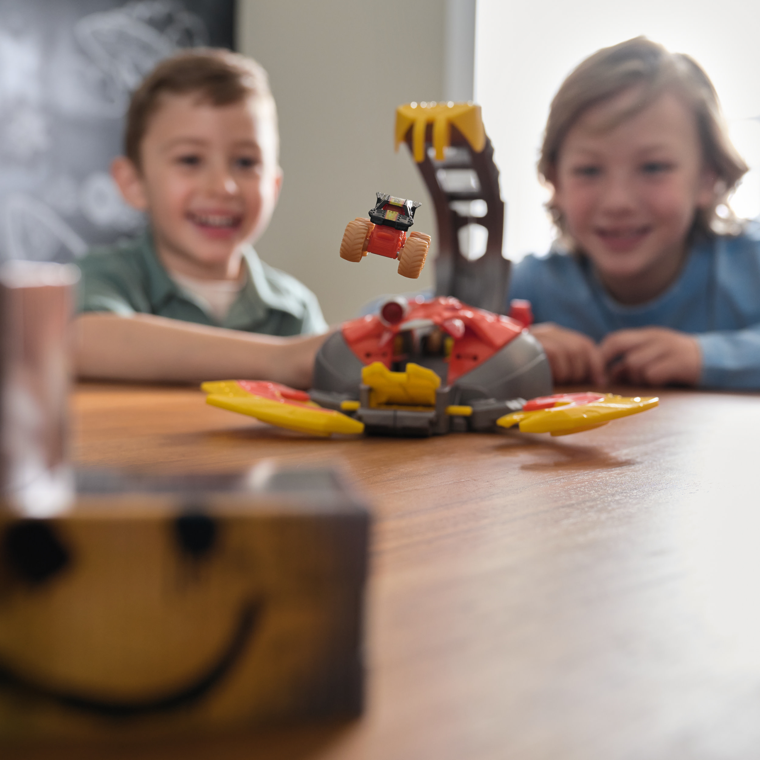 Dwóch chłopców uśmiecha się do zabawki Monster Jam, z ciężarówką w powietrzu. Uśmiechnięta buzia jest na drewnianym klocku.