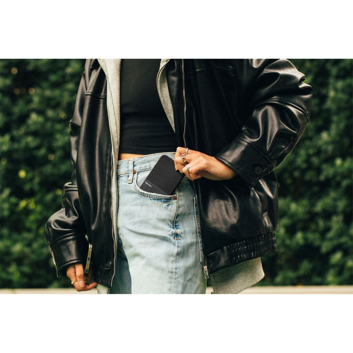 Une personne en veste noire et jean met un appareil noir dans sa poche, avec un fond vert.