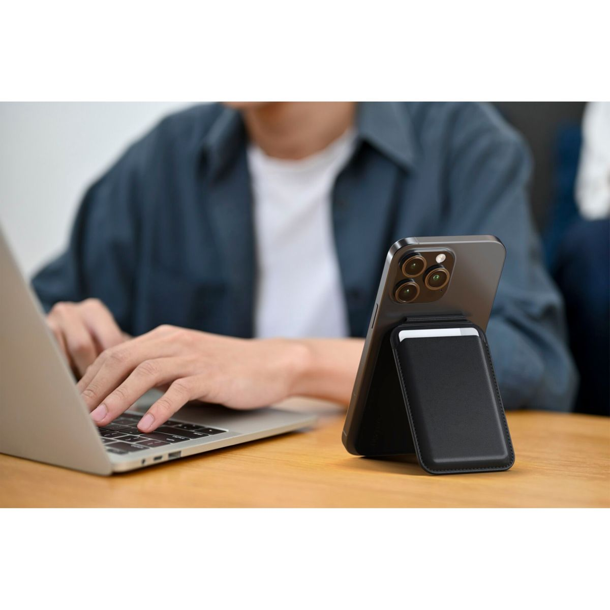 Un telefono con un porta carte su una scrivania di legno. Una persona usa un laptop sullo sfondo.