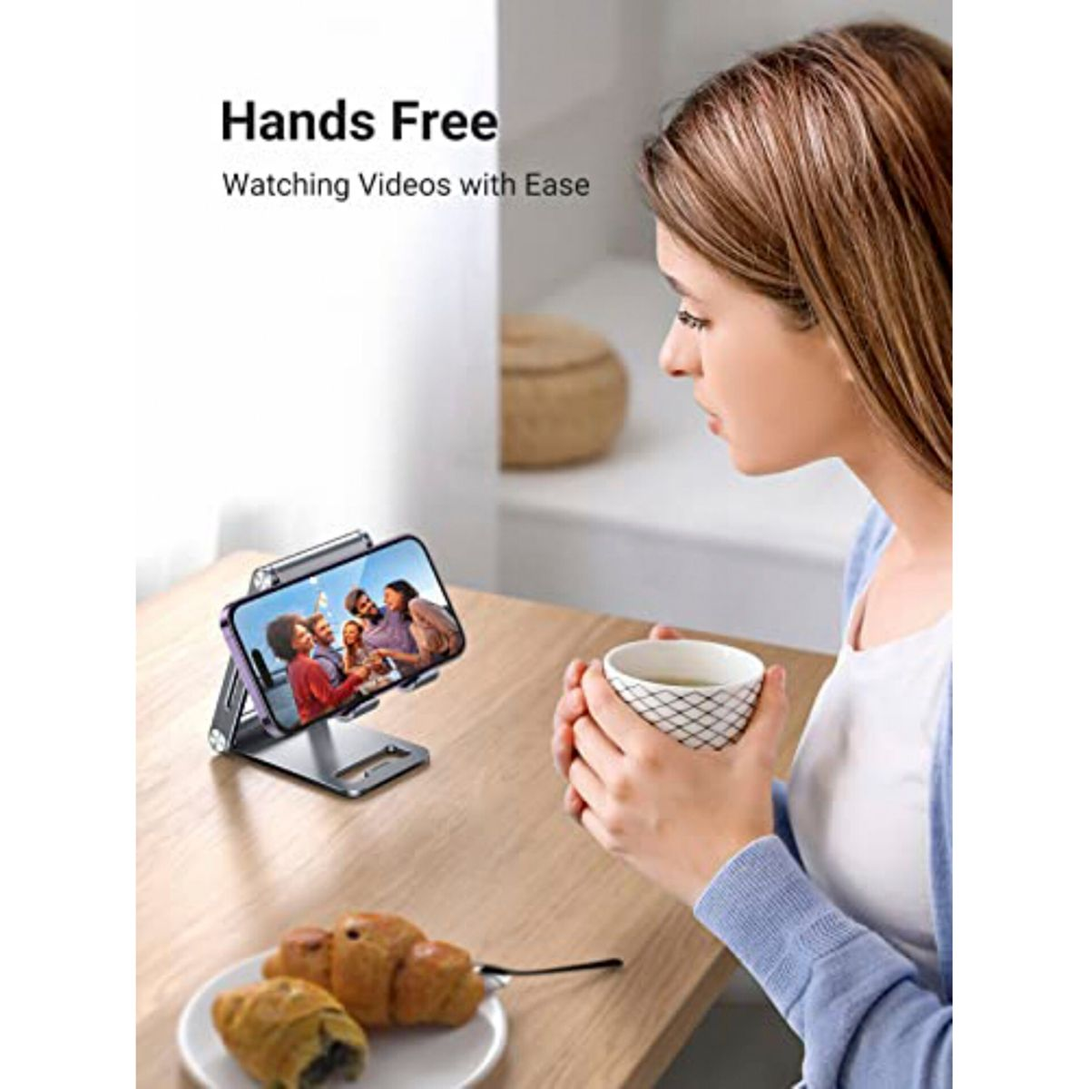 Eine Frau schaut ein Video auf ihrem Telefon, das auf einem Ständer steht. Croissants liegen auf einem Teller auf dem Tisch.