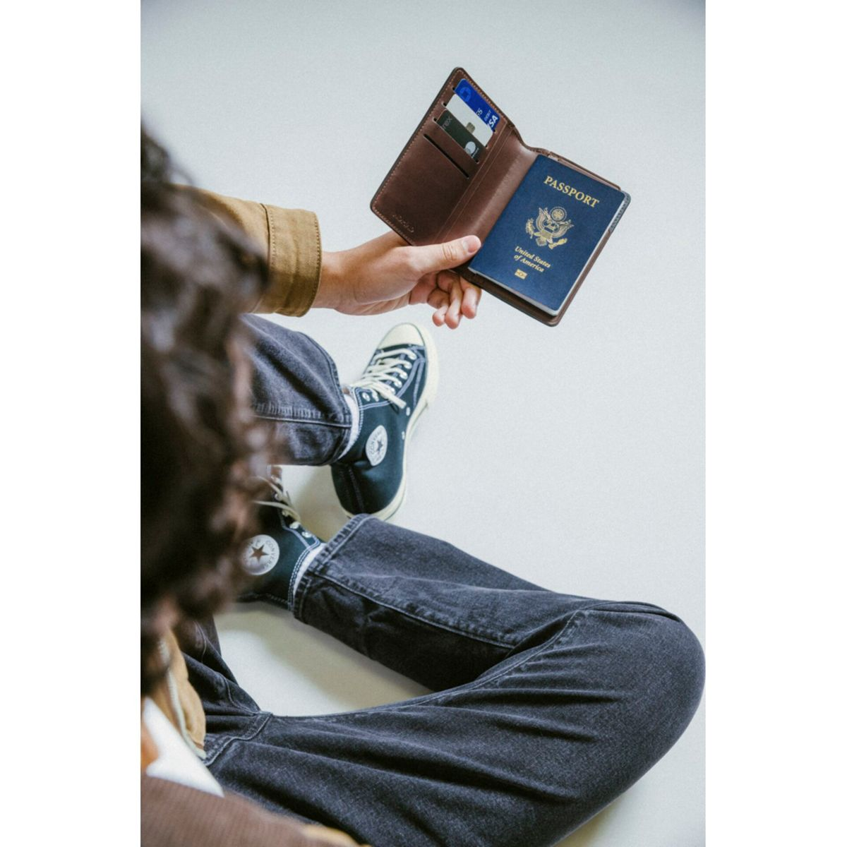 Una persona tiene in mano un passaporto in un portafoglio di pelle marrone, aperto sulla pagina del passaporto. Scarpe Converse visibili.