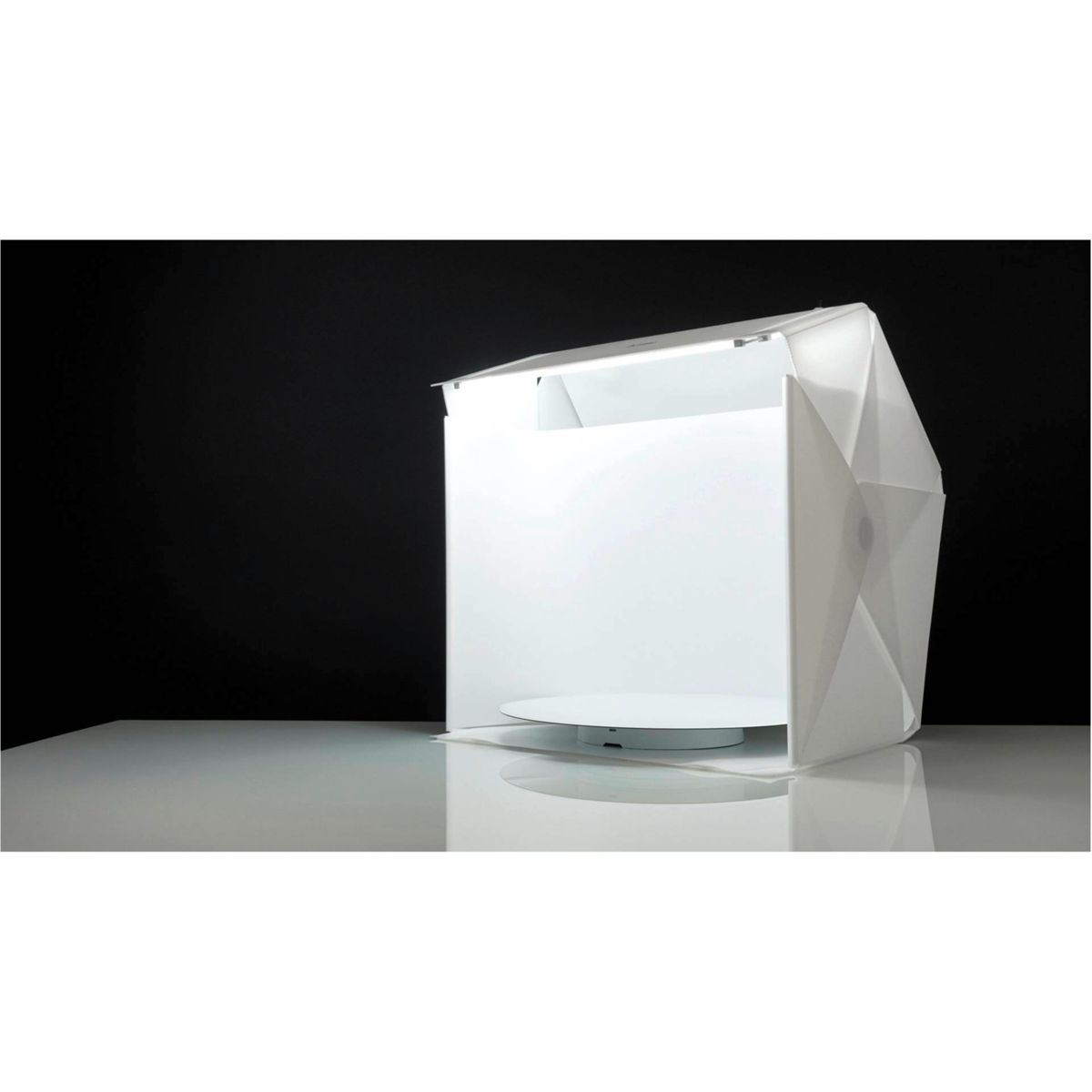 Una caja de estudio fotográfico blanca sobre una superficie blanca, con fondo negro. La caja tiene una plataforma circular blanca.