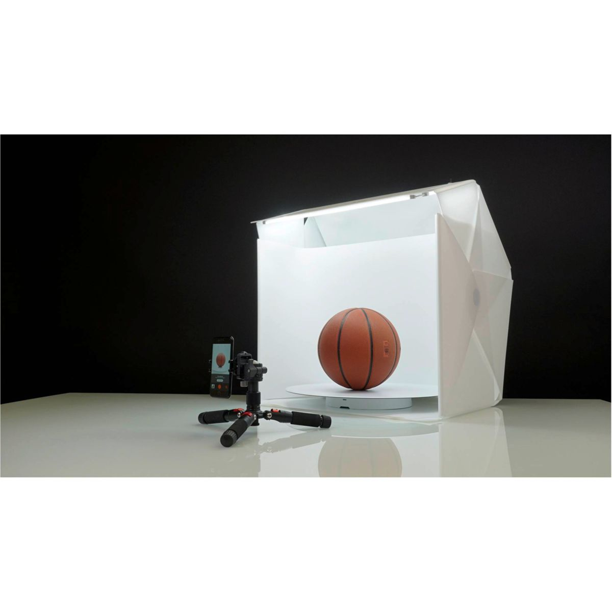 Un balón de baloncesto dentro de una caja de estudio fotográfico blanca. Hay un teléfono y un trípode a la izquierda. El fondo es negro.