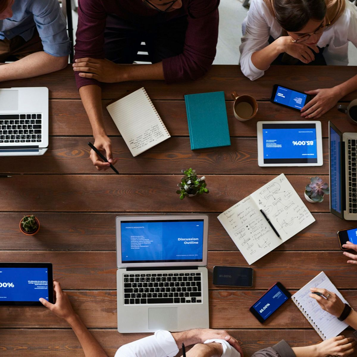 Widok z góry na drewniany stół z kilkoma osobami, laptopami, tabletami i notatnikami. Przedmioty są ułożone na stole.
