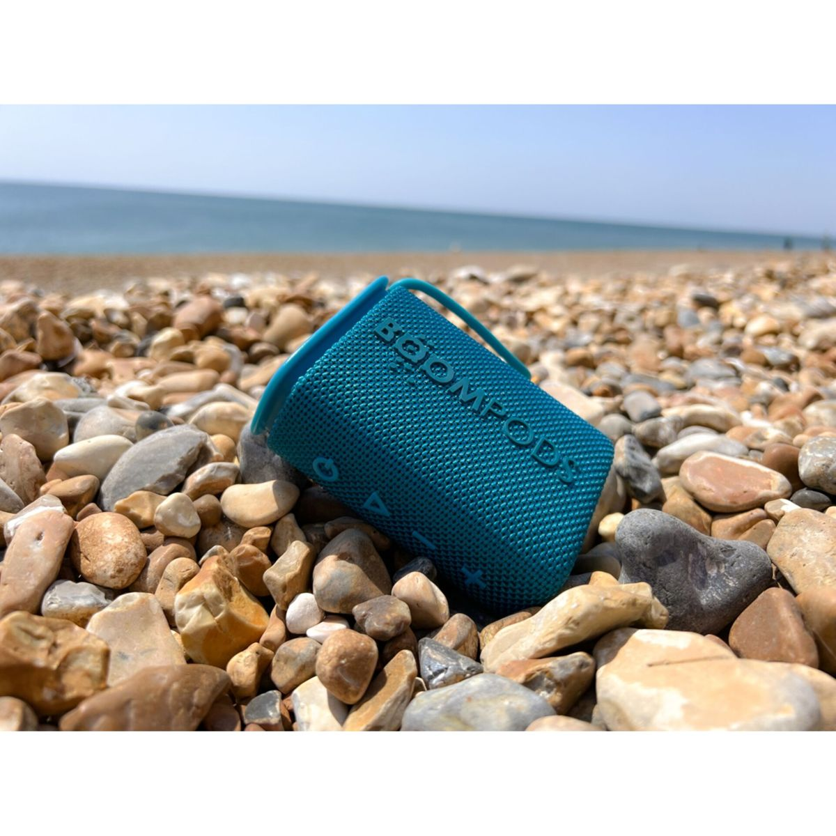 Türkisfarbener tragbarer Lautsprecher an einem Kiesstrand. Blauer Himmel im Hintergrund.