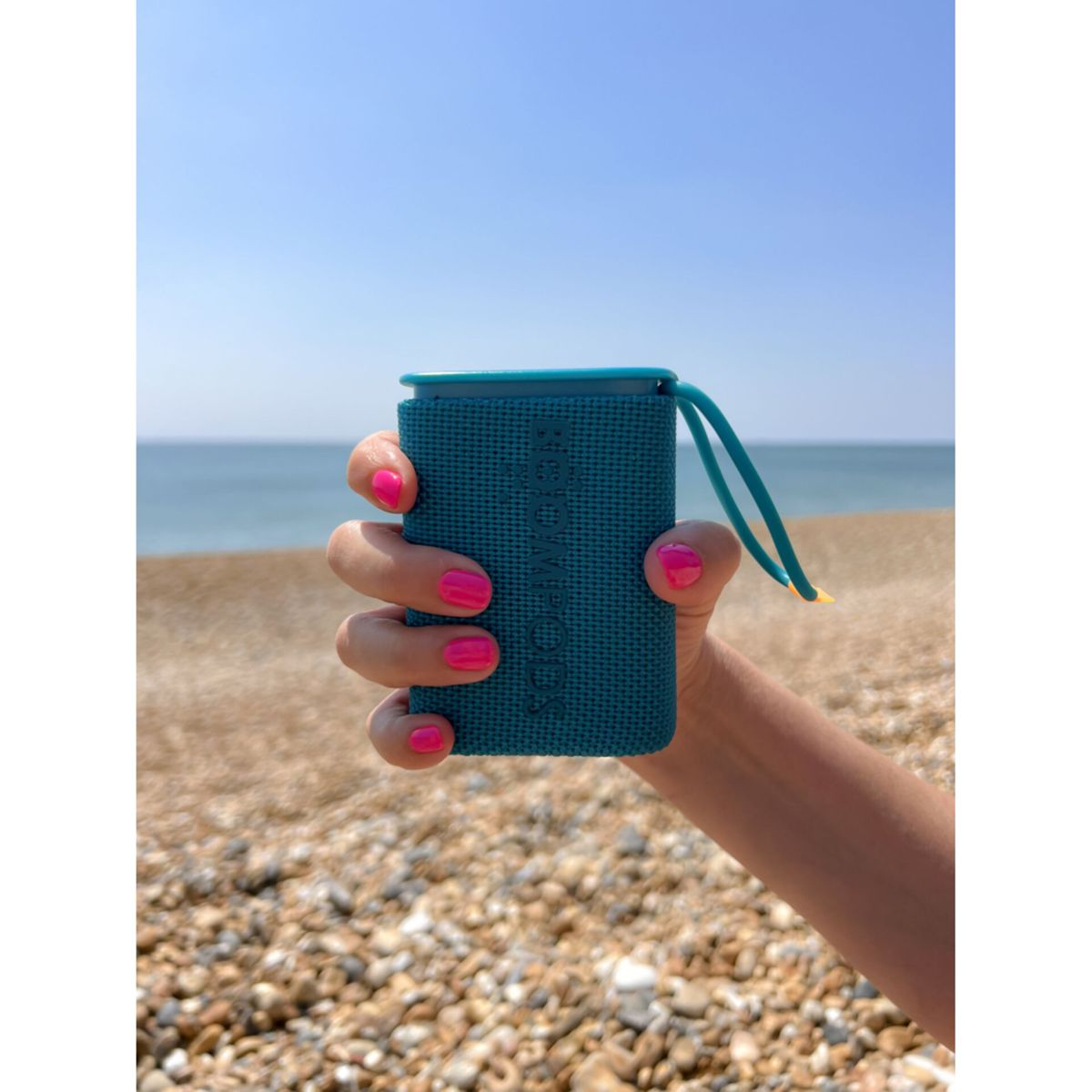 Hand hält einen türkisfarbenen tragbaren Lautsprecher an einem Kiesstrand, rosa Nägel.