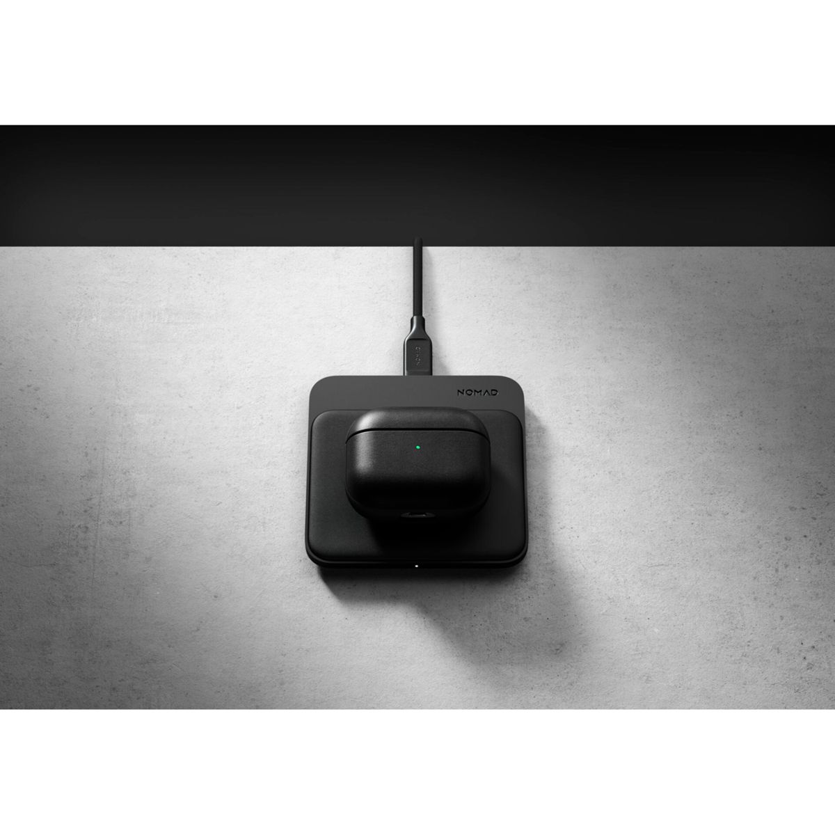 Custodia AirPods nera su un pad di ricarica, collegata tramite un cavo a una parete. Il pad e la parete sono su una superficie grigia.