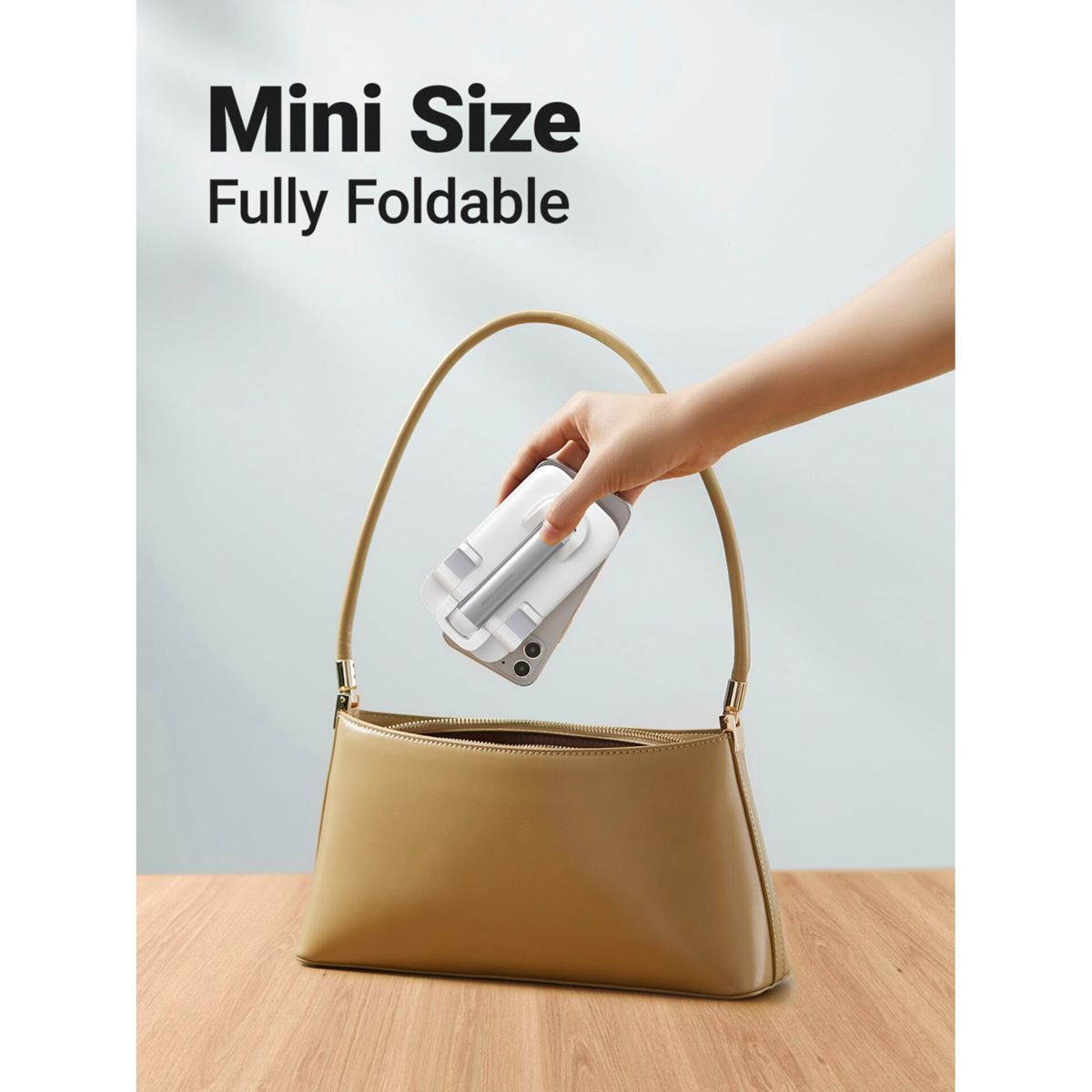 Ręka trzymająca składany stojak na telefon w beżowej torebce. Tekst mówi: „Mini rozmiar, w pełni składany”.