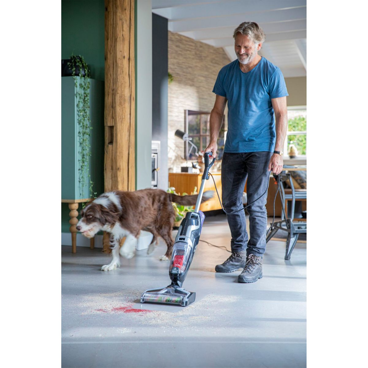 Mann reinigt den Boden mit einem Bodenreiniger. Im Hintergrund läuft ein Hund.