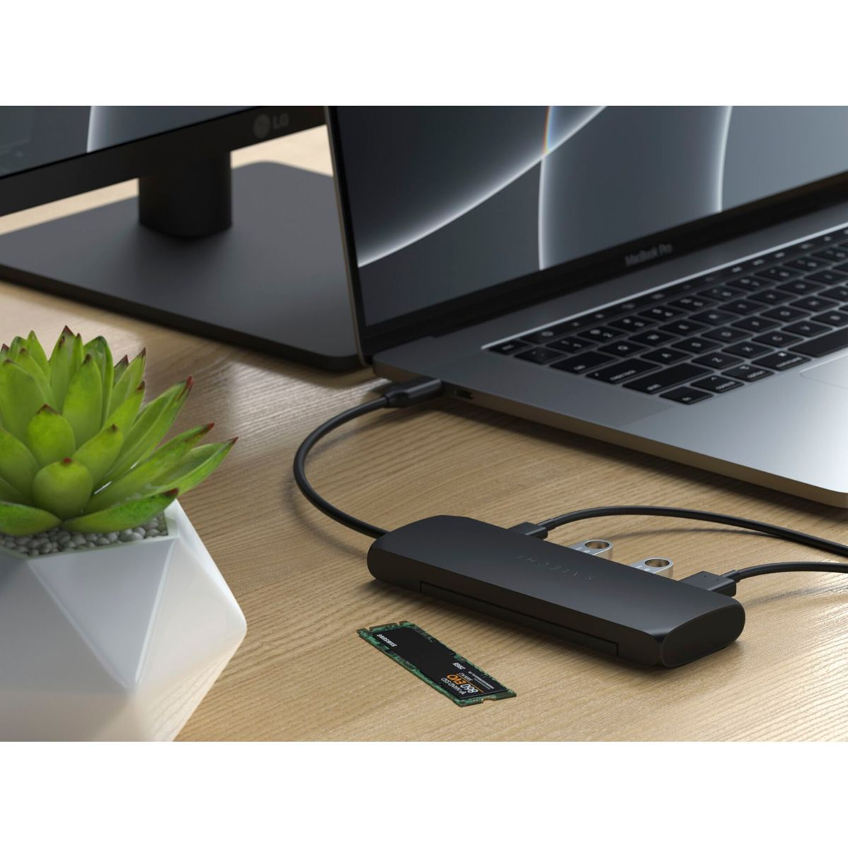 Ein Laptop, ein Monitor und ein Hub sind auf einem Holzschreibtisch angeschlossen. Neben dem Monitor steht eine grüne Pflanze.