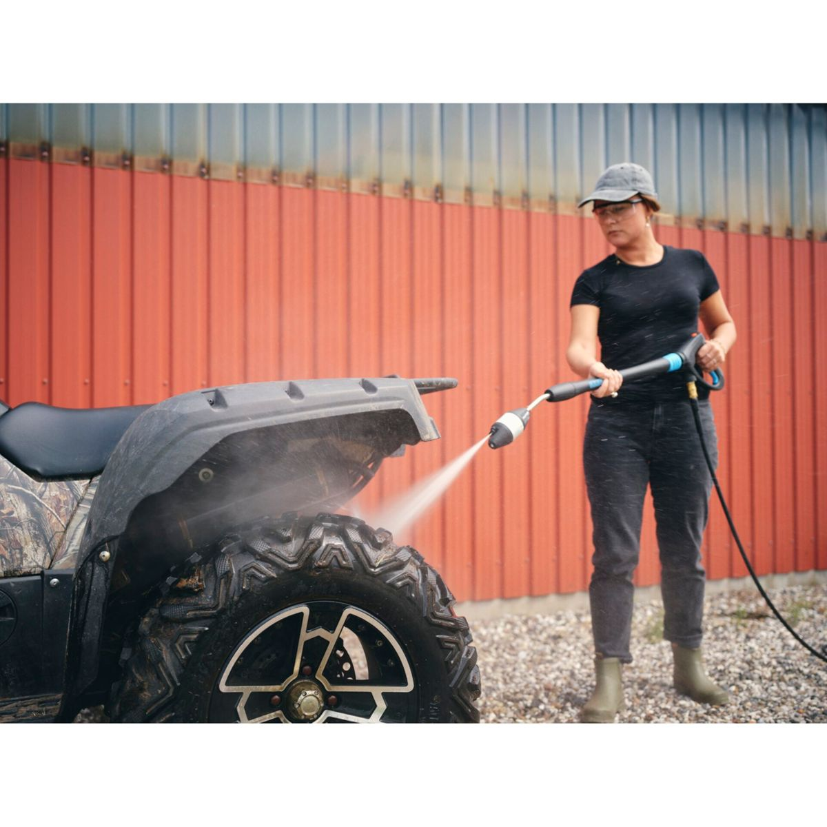 Une femme lave un VTT avec un nettoyeur haute pression devant un bâtiment rouge par temps nuageux.