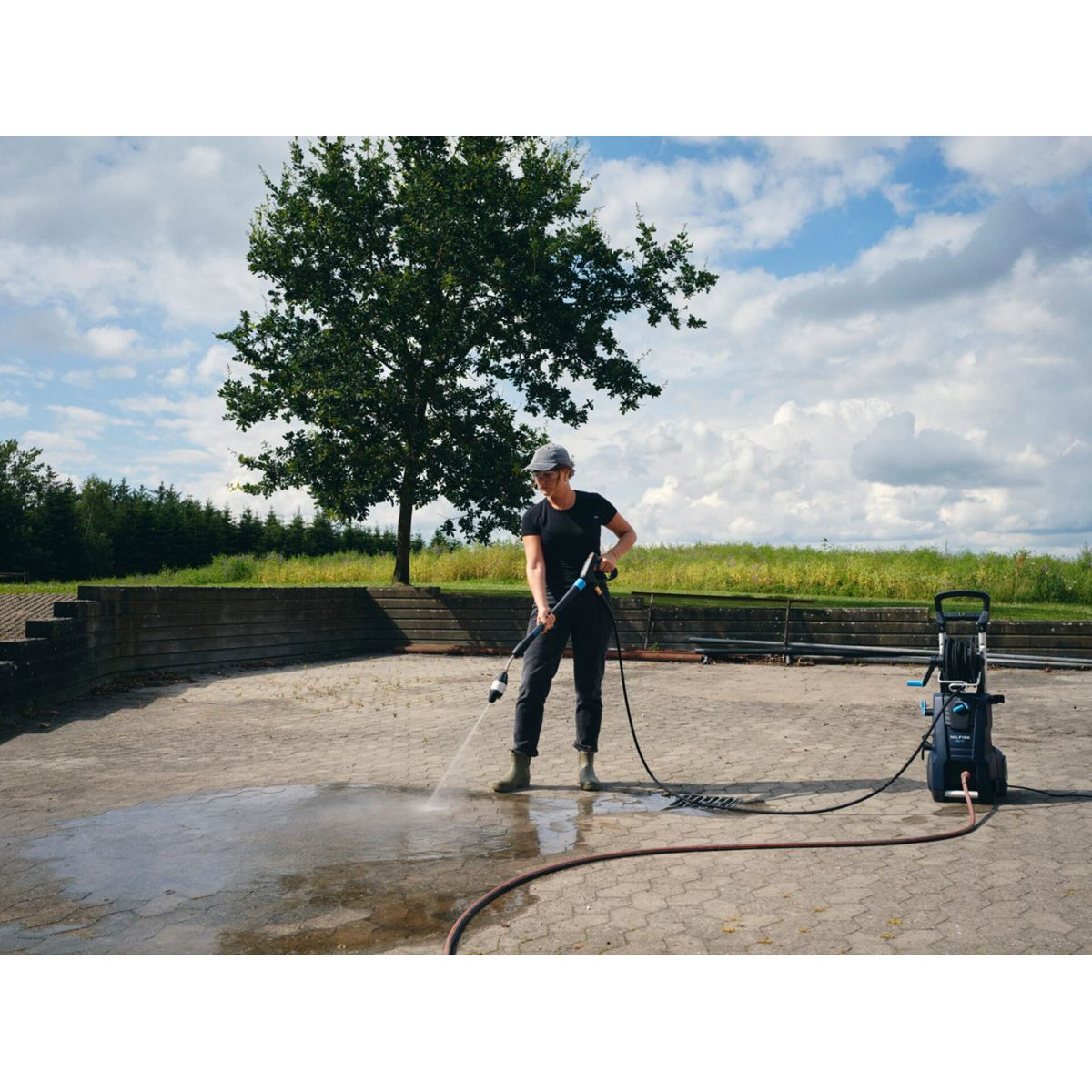 Une femme lave une surface en béton avec un nettoyeur haute pression par une journée ensoleillée.