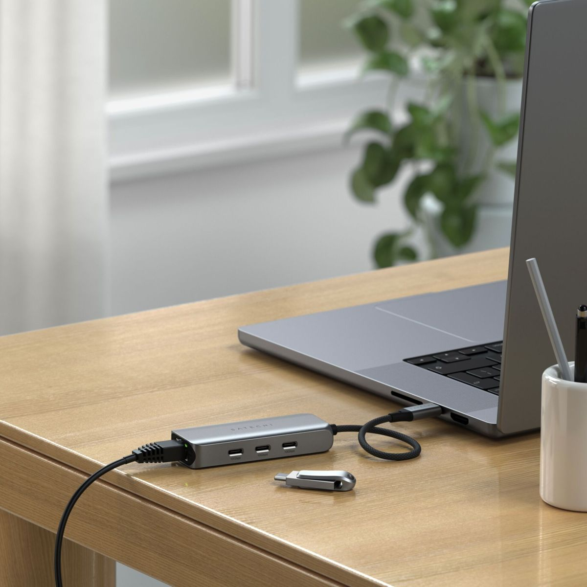 Ein Laptop, der mit einem silbernen Hub auf einem Holztisch verbunden ist, mit einer Pflanze im Hintergrund.
