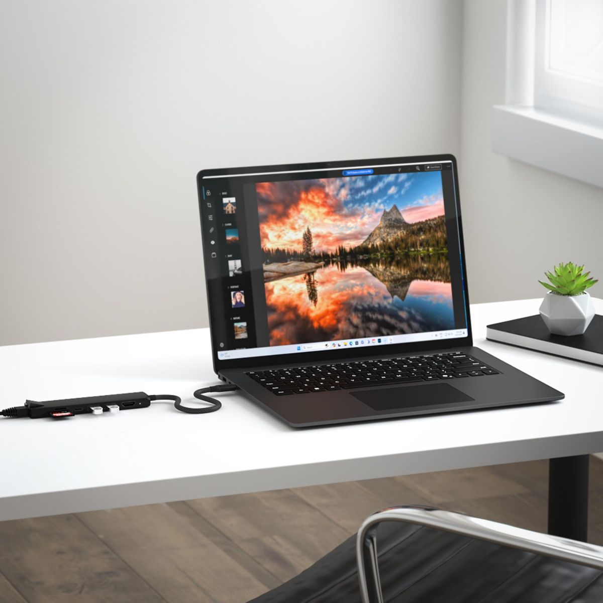 Laptop auf einem weißen Schreibtisch. Auf dem Bildschirm ist ein Landschaftsfoto zu sehen. Ein Hub ist angeschlossen.