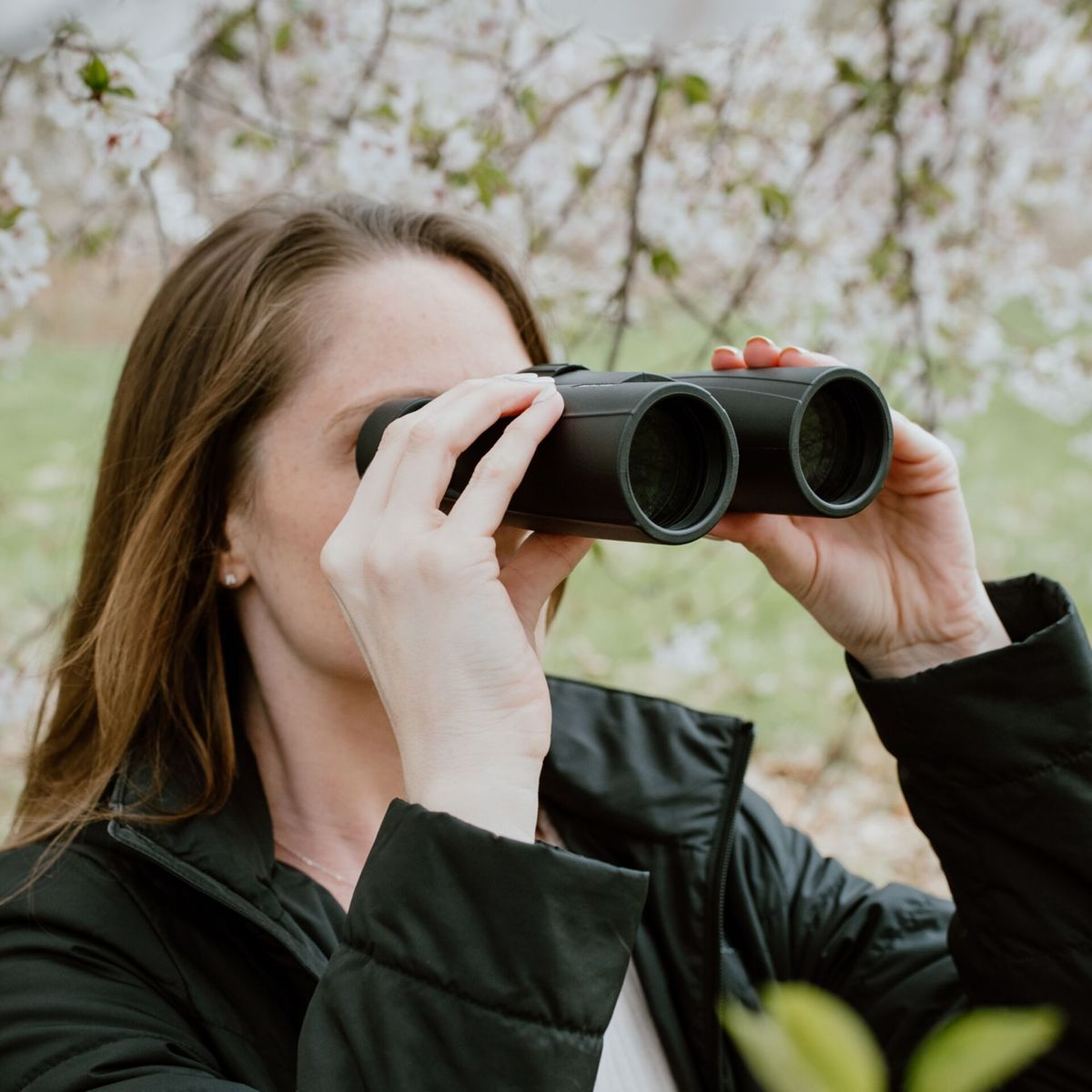 Donna che guarda attraverso un binocolo nero, sfondo sfocato di fiori. Indossa una giacca nera.
