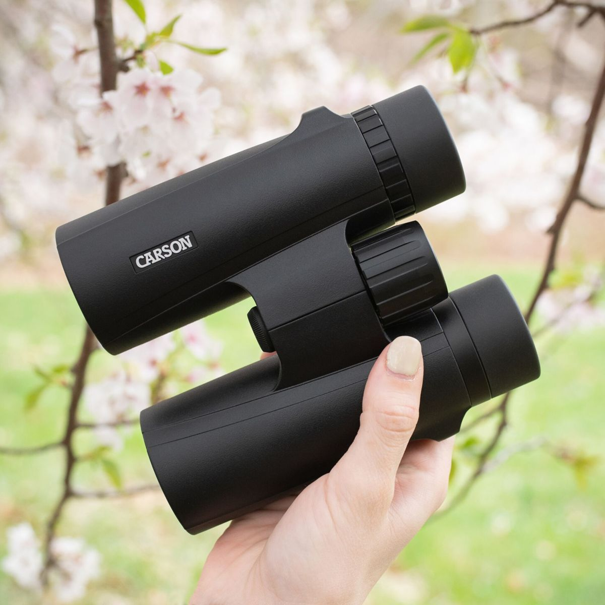 Binocolo nero tenuto in mano. Testo bianco 'CARSON' sul lato. Sfondo floreale sfocato.