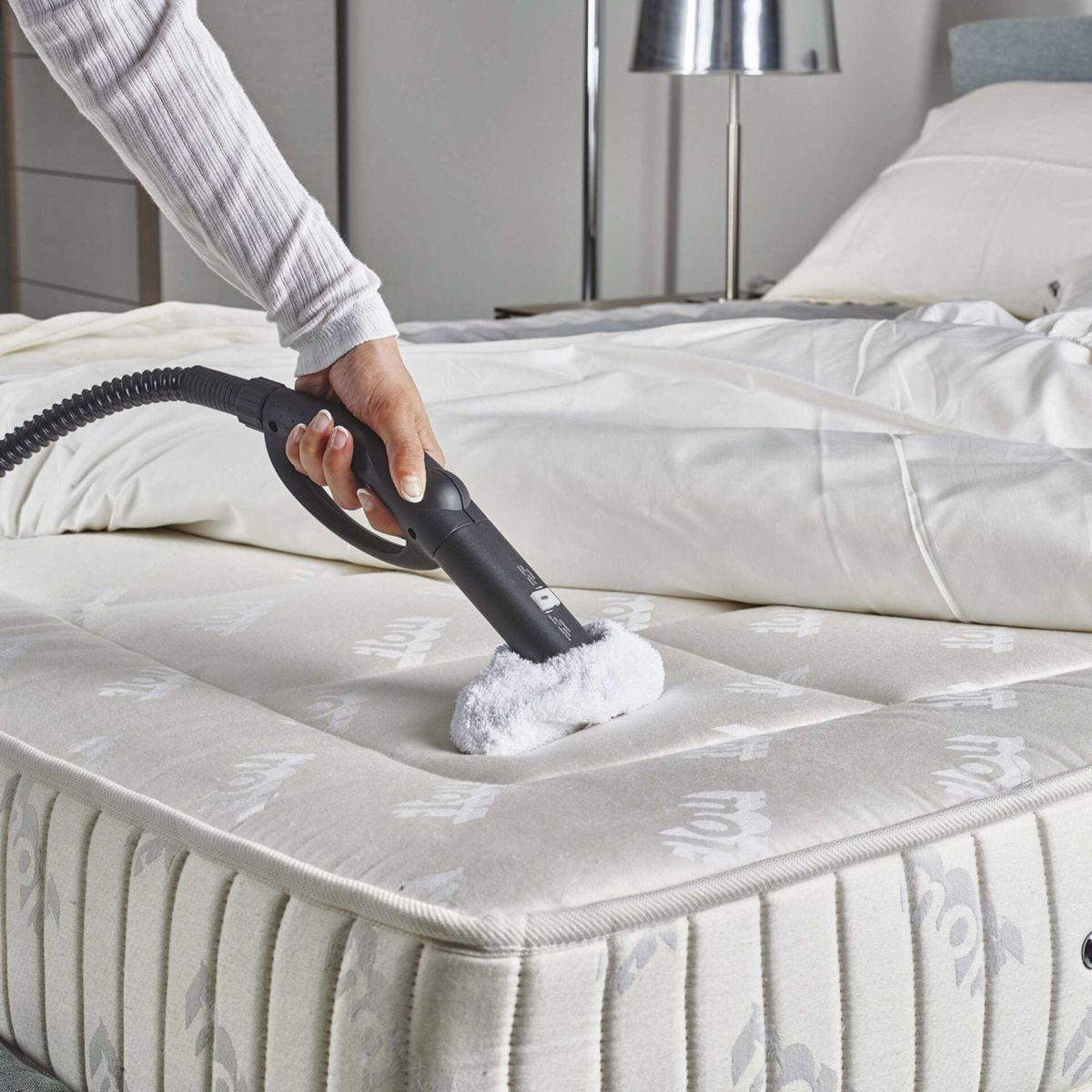 Una persona limpiando un colchón con un vaporizador de mano. Cama blanca y sábana en el fondo.