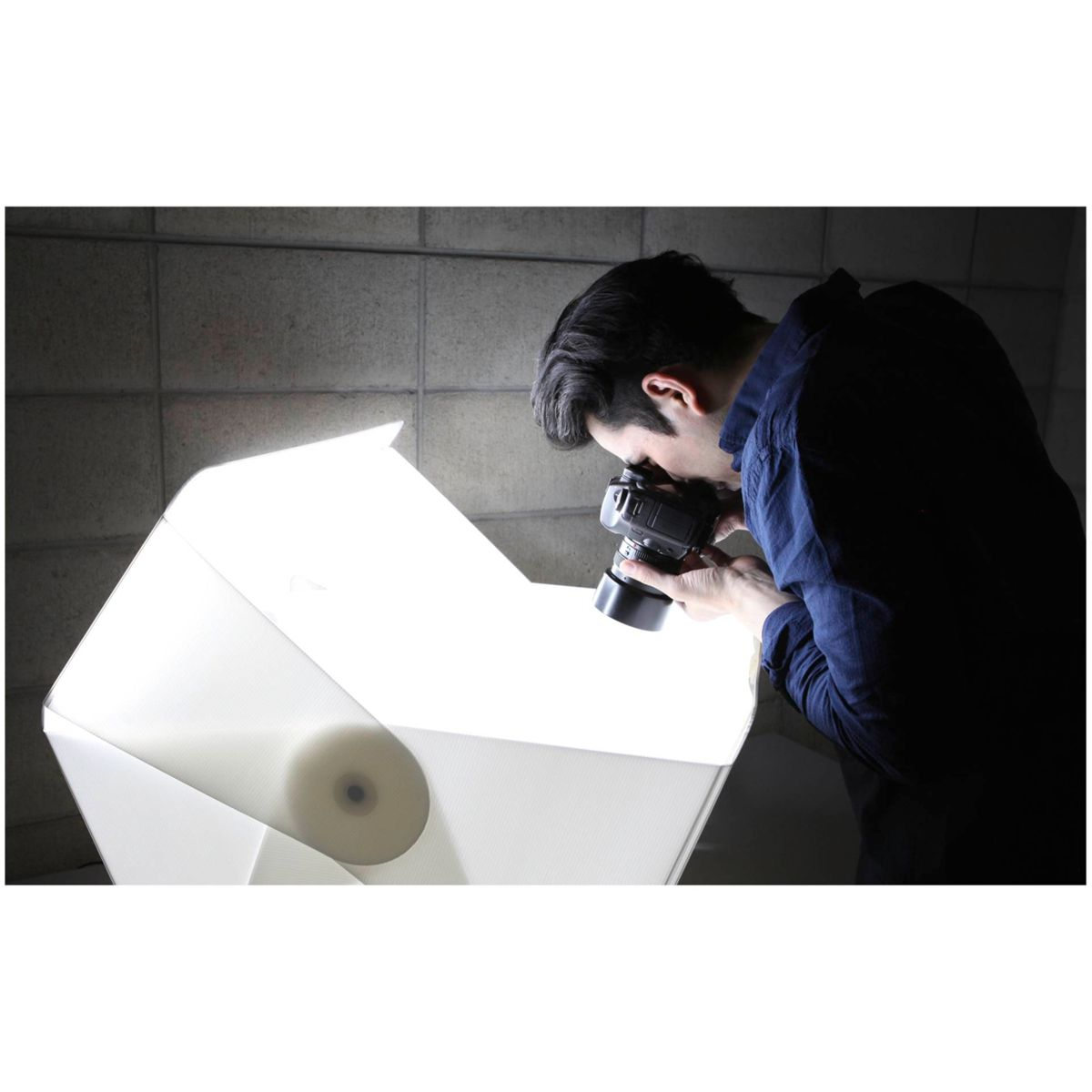 Eine Person fotografiert einen Gegenstand in einer weißen Lichtbox vor einer grauen Wand.
