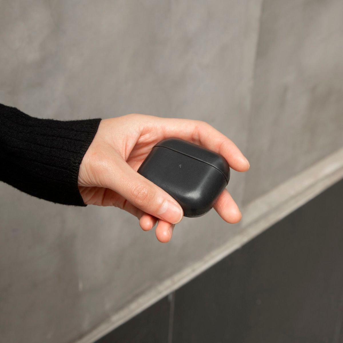 Une main tient un étui AirPods noir, avec un mur texturé en arrière-plan.