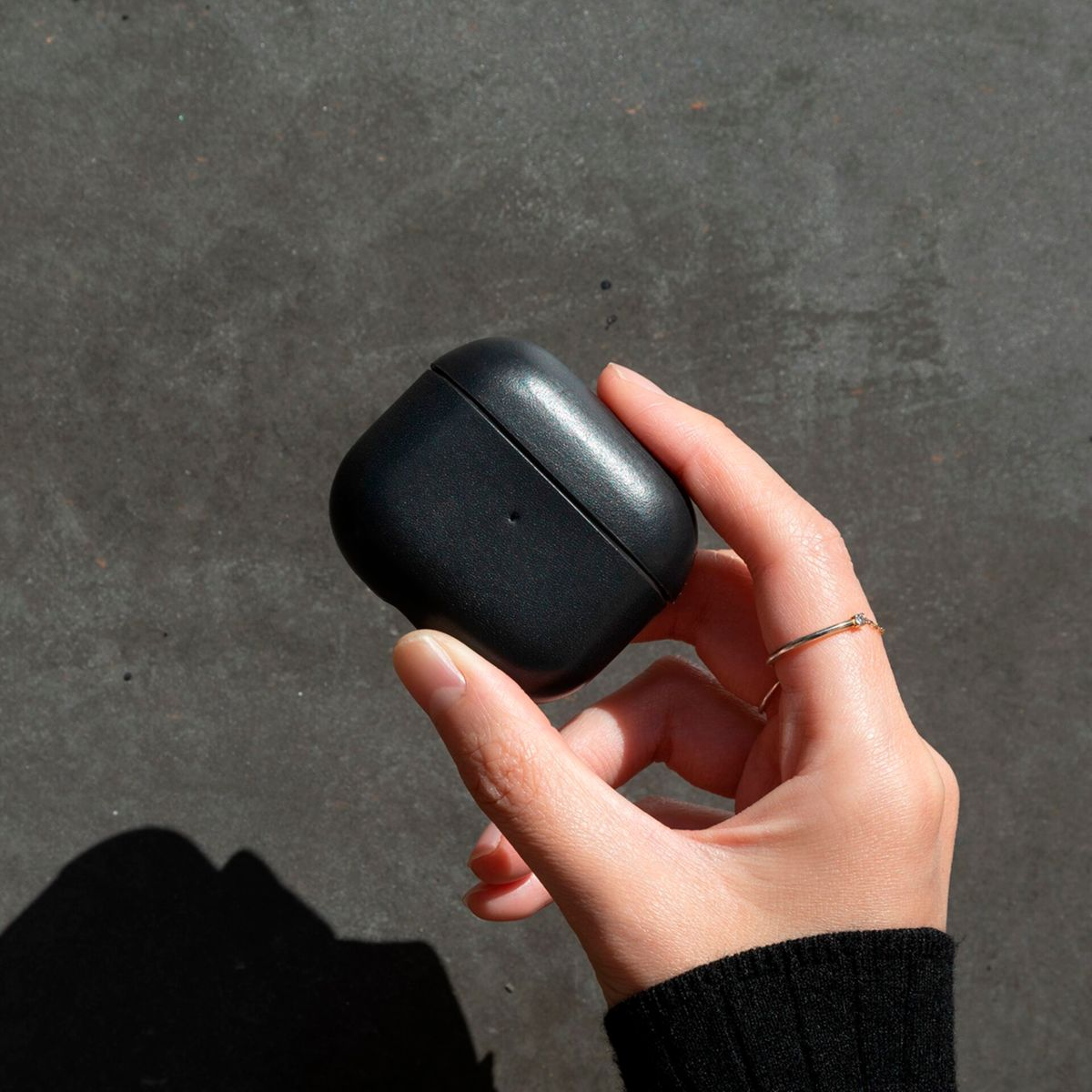 Une main tient un étui AirPods noir sur un fond gris texturé.