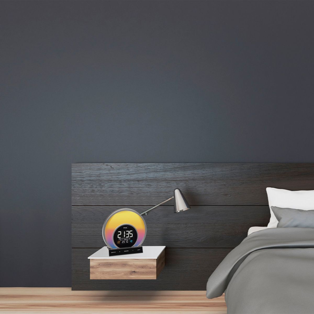 Wecker auf einem Nachttisch mit einem Bett im Hintergrund. Die Uhr zeigt Zeit und Datum an.