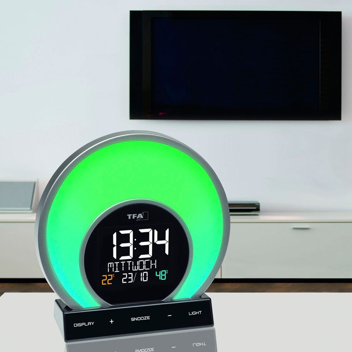 Ein Wecker mit grüner Lichtanzeige auf einem Glastisch, mit einem Fernseher im Hintergrund.