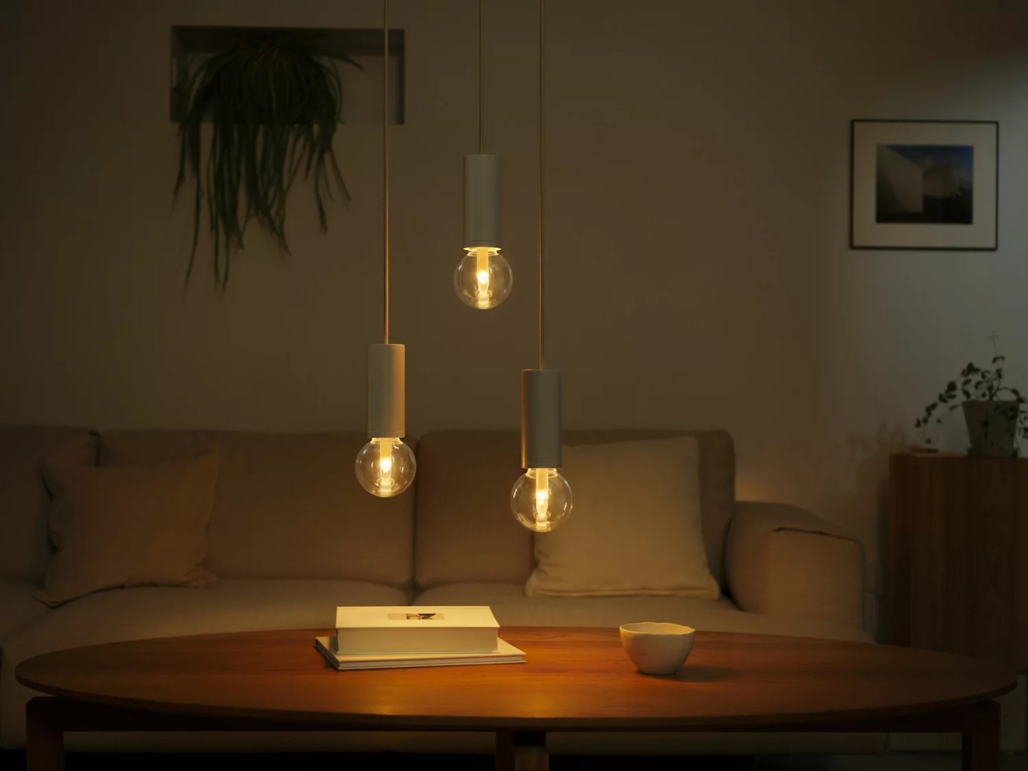 Trzy wiszące lampy z żarówkami nad okrągłym stołem, z sofą i roślinami w tle.