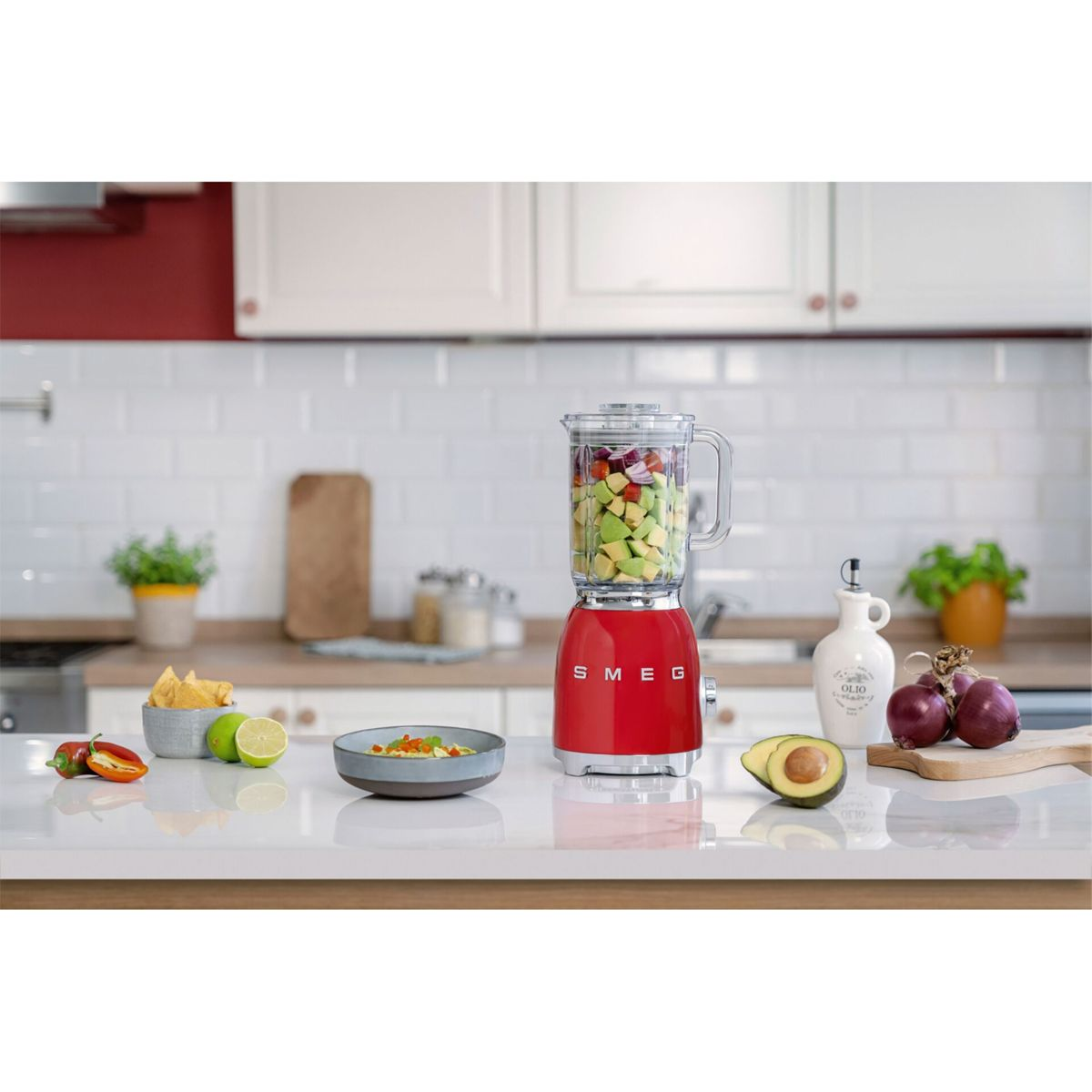 Czerwony blender wypełniony pokrojonymi owocami i warzywami stoi na białym blacie z artykułami spożywczymi.