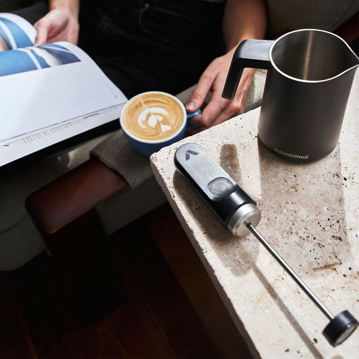 Eine Person rührt Kaffee mit einem Milchaufschäumer um. Eine Tasse Kaffee mit Latte Art und ein schwarzer Milchkrug sind daneben.