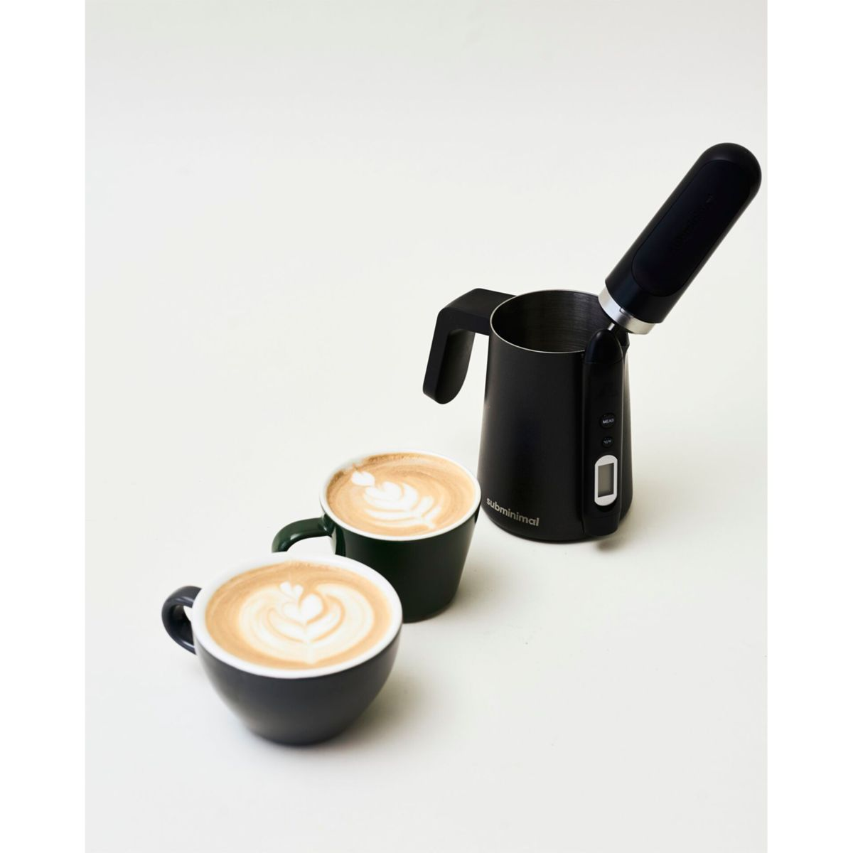 Zwei Tassen Kaffee mit Latte Art und ein schwarzer Milchaufschäumer auf weißer Oberfläche.