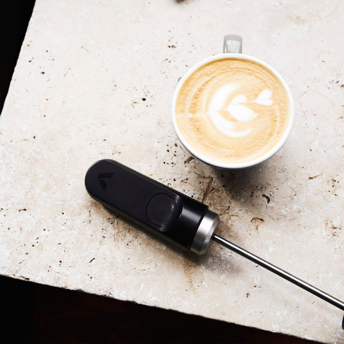 Eine Tasse Kaffee mit Latte Art neben einem schwarzen Milchaufschäumer auf einer Steinoberfläche. Der Aufschäumer ist inaktiv.