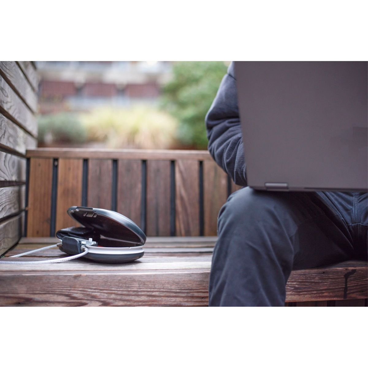 Una persona sentada en un banco de madera, usando una laptop. Una caja fuerte con cable está junto a ellos.