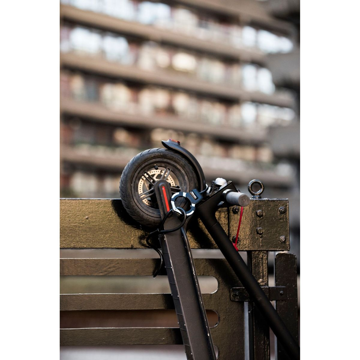 Scooter nero parcheggiato vicino a una recinzione con un lucchetto, edificio sfocato sullo sfondo.