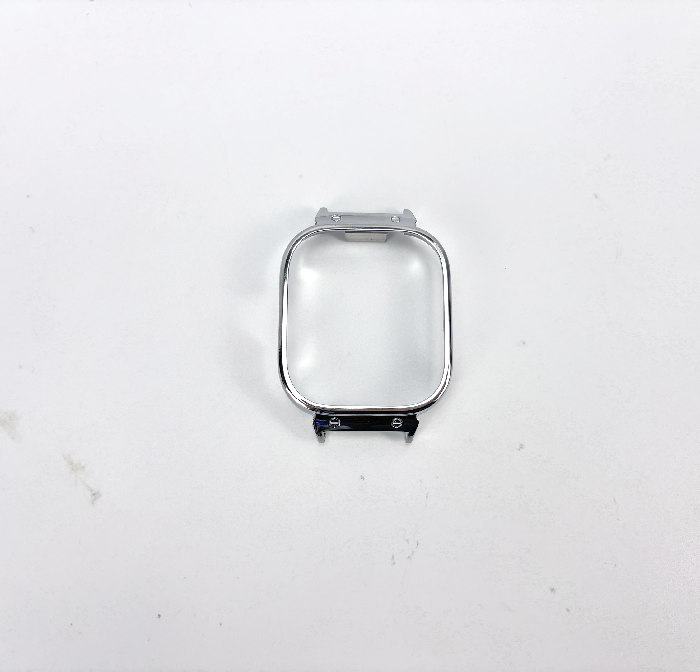 Srebrna, prostokątna ramka zegarka z zakrzywioną górą. Leży na białej powierzchni.