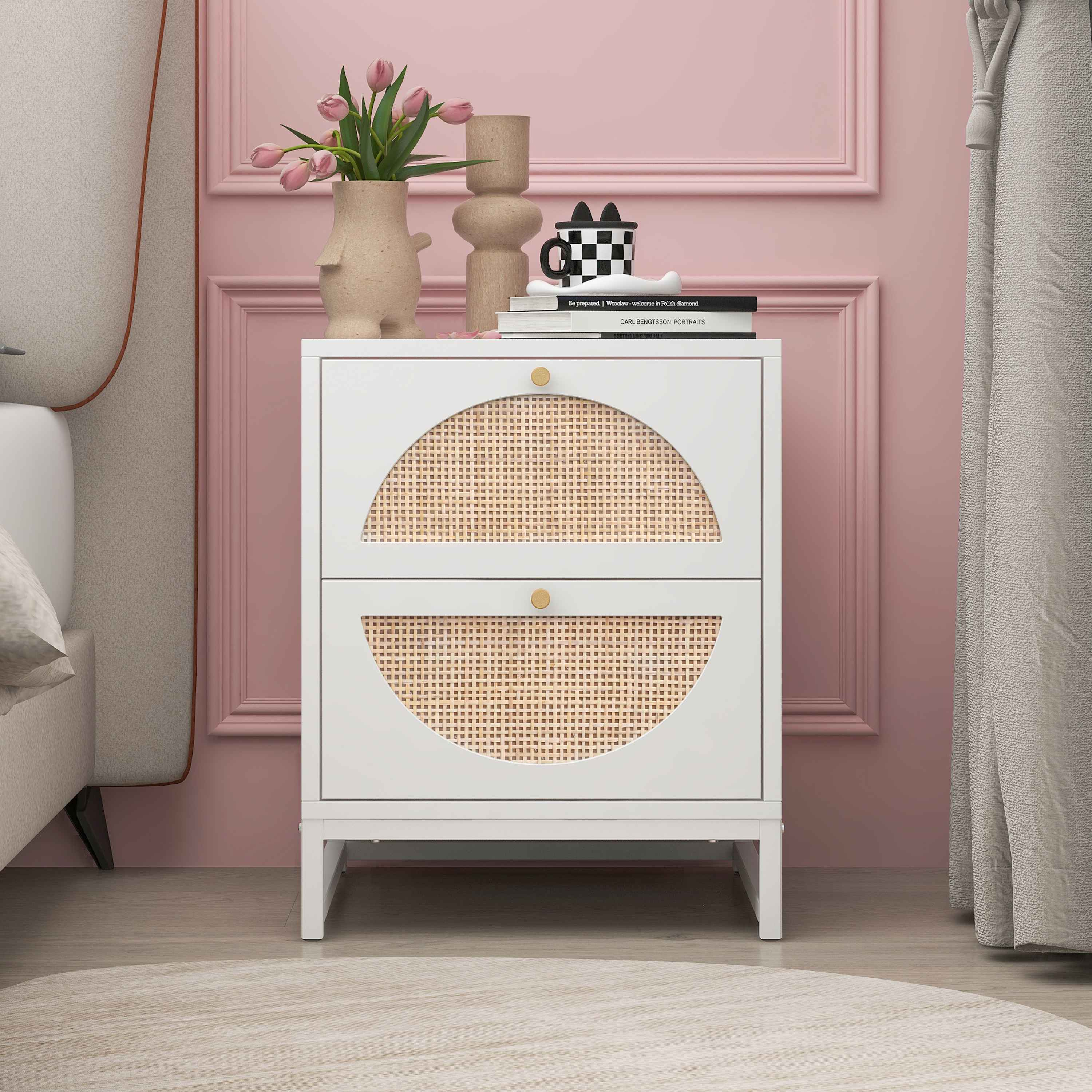 Weißer Nachttisch mit zwei Schubladen und Rattandesign, goldfarbene Griffe, an einer rosa Wand.