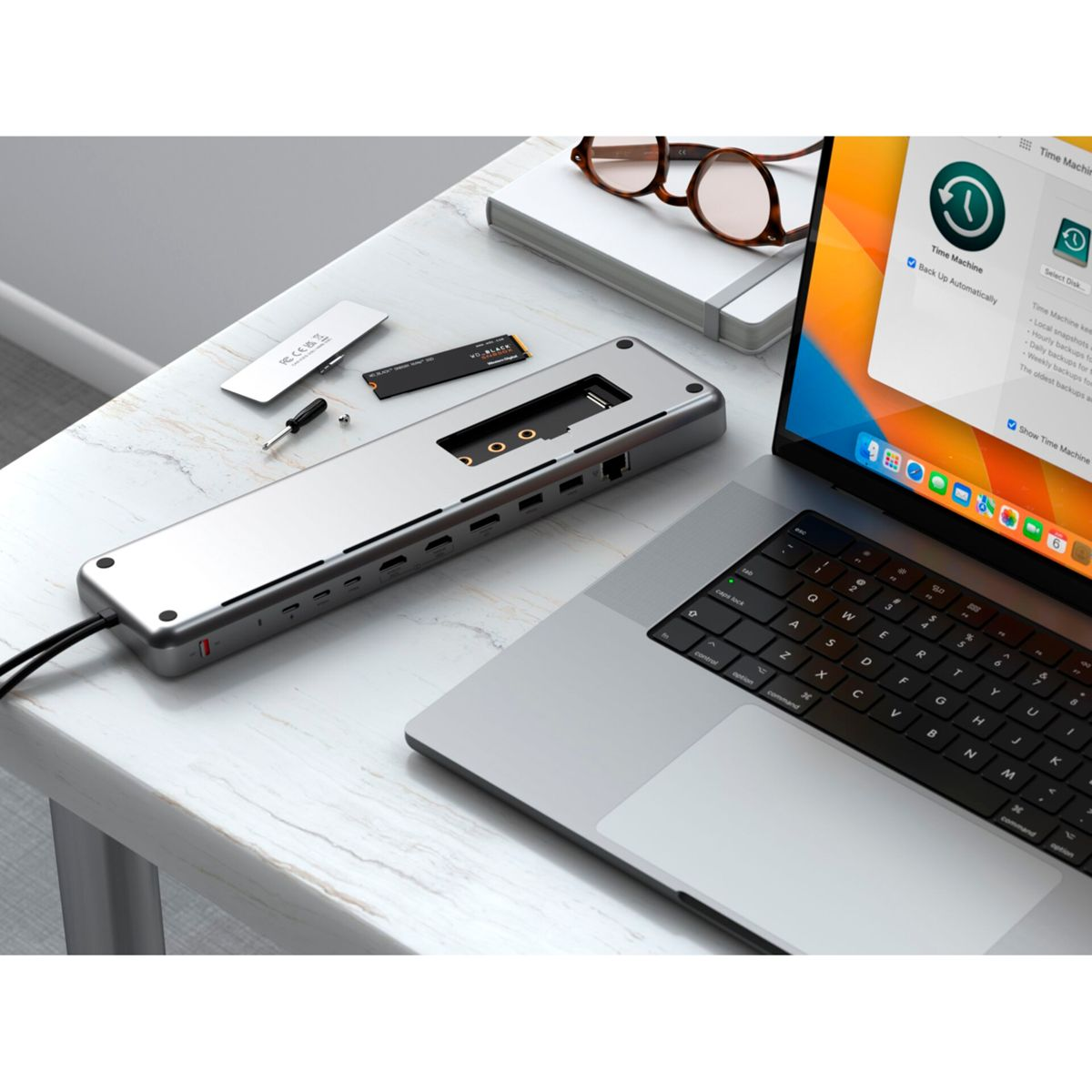 Un hub argentato si collega a un laptop. Altri dispositivi, come un disco rigido, sono vicini.