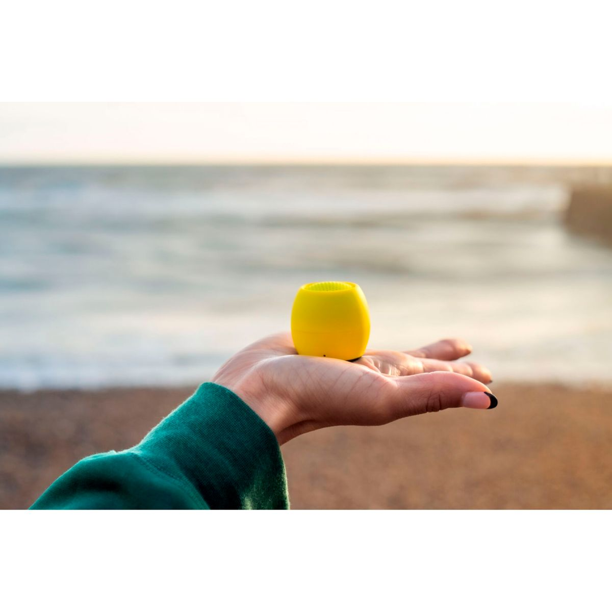 Una mano tiene un piccolo altoparlante giallo su uno sfondo sfocato di mare e tramonto.
