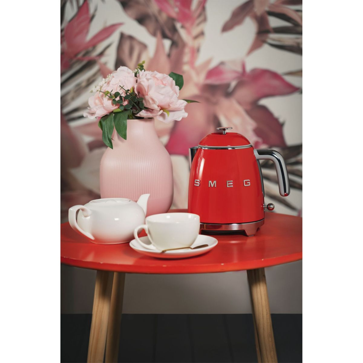 Roter SMEG-Wasserkocher auf einem roten Tisch mit weißer Teekanne, Tasse und rosa Blumen in einer Vase.