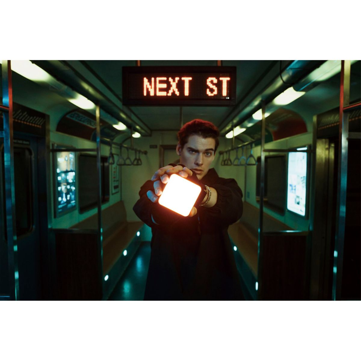 Ein Mann in einer U-Bahn hält ein quadratisches Licht zur Kamera. Ein Schild liest 'NEXT ST'.