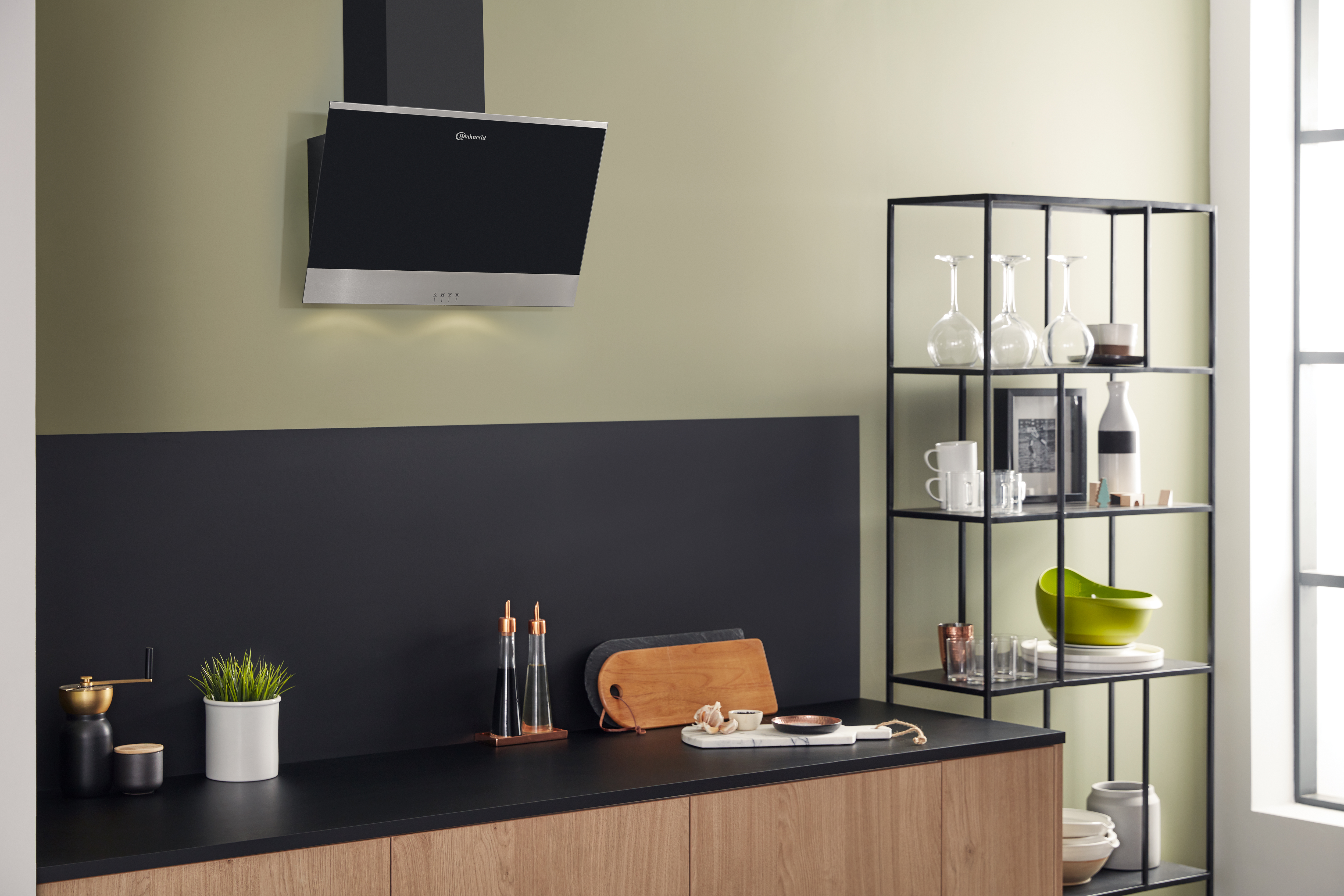 Cucina moderna con cappa nera, piano di lavoro nero e mobili in legno. Una mensola contiene bicchieri e decorazioni.