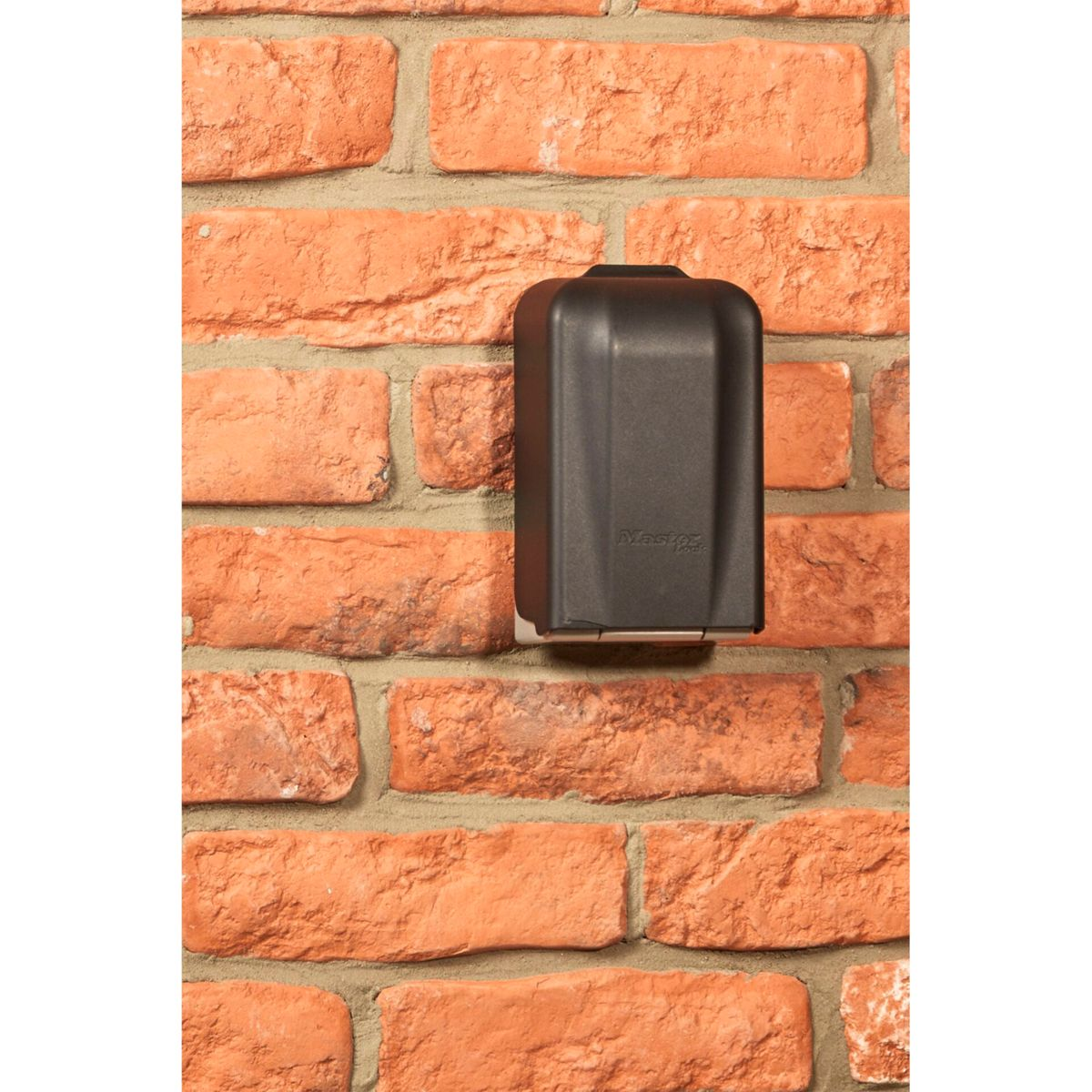 Ein schwarzer Master Lock Schlüsseltresor ist an einer roten Backsteinmauer befestigt. Er ist geschlossen und rechteckig.