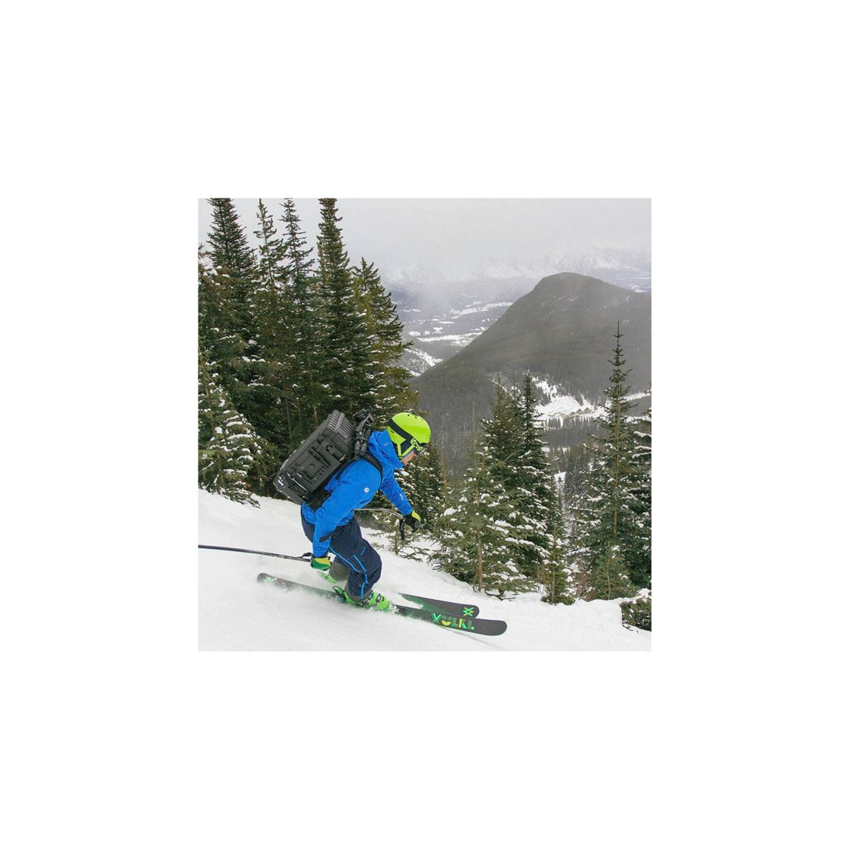 Ein Skifahrer fährt einen verschneiten Berg hinunter. Der Skifahrer trägt Blau, Grün und einen Helm. Bäume sind an den Seiten.