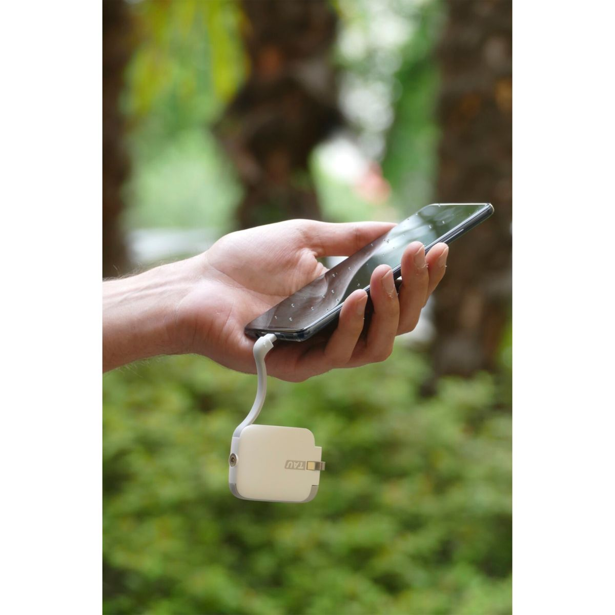 Une main tenant un téléphone connecté à un chargeur portable blanc avec 'TAU' dessus, dans un cadre extérieur flou.