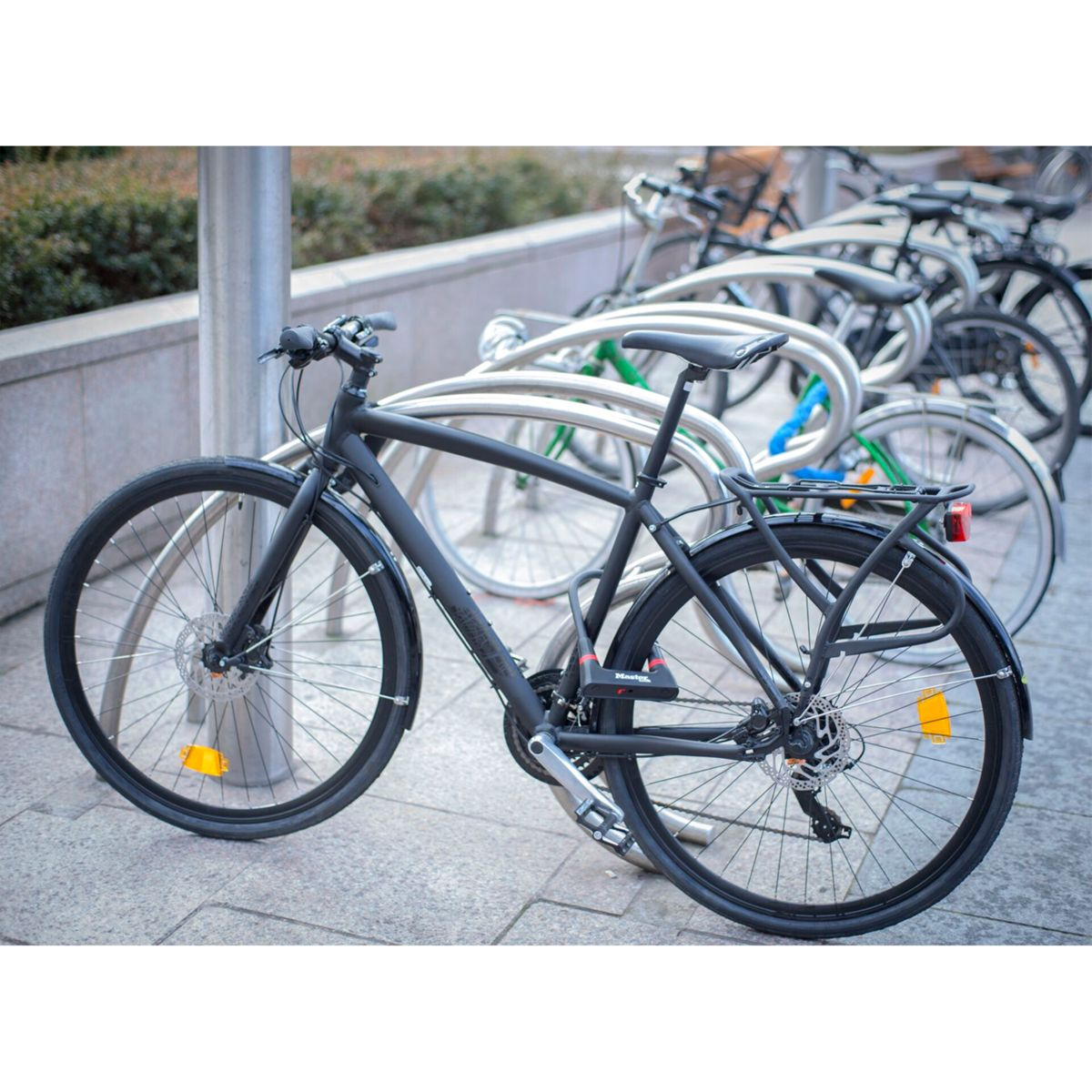 Schwarzes Fahrrad an einem Ständer angeschlossen, andere Fahrräder im Hintergrund, draußen.