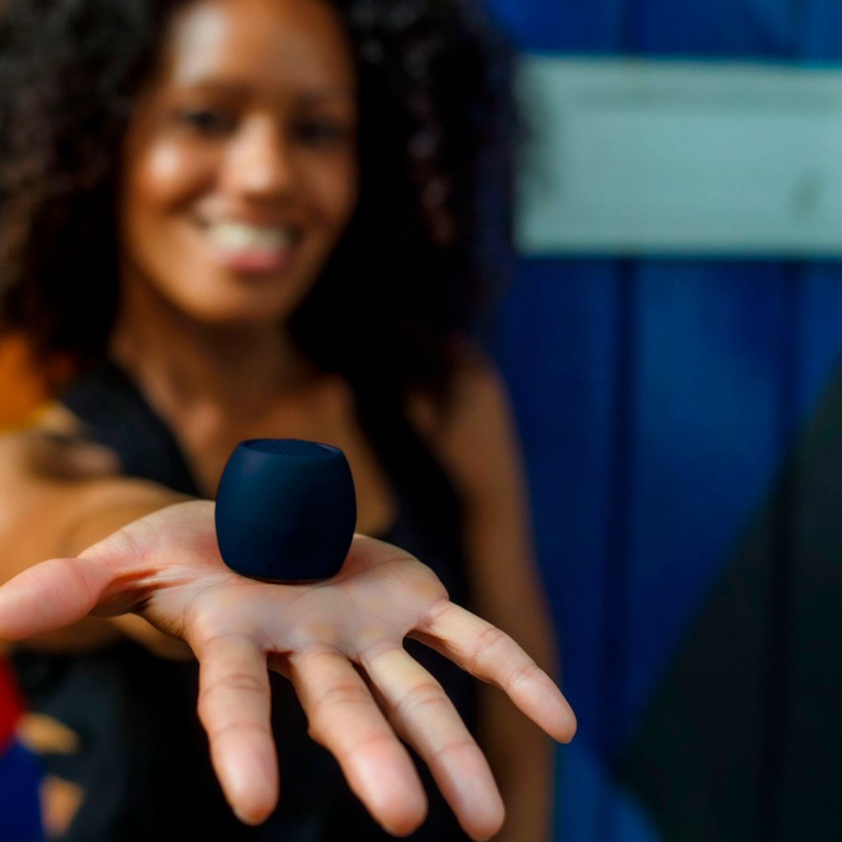Kobieta trzymająca mały, ciemnoniebieski głośnik na otwartej dłoni, uśmiechając się.