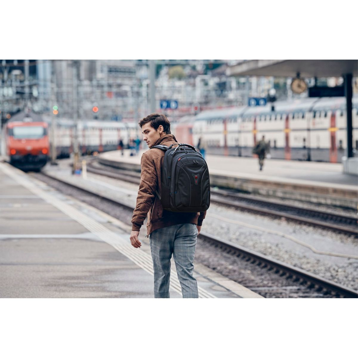 Mann mit Rucksack am Bahnhof, Zug im Hintergrund. Braune Jacke, graue Hose.