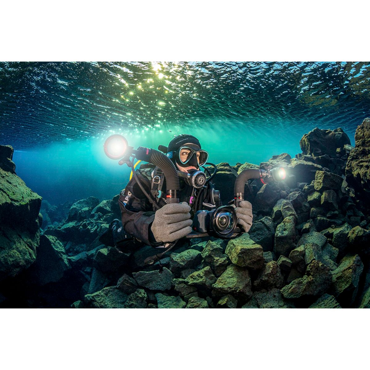 Ein Taucher in einer Unterwasserhöhle. Hält eine Kamera mit zwei Lichtern. Felsen und Wasser sichtbar.