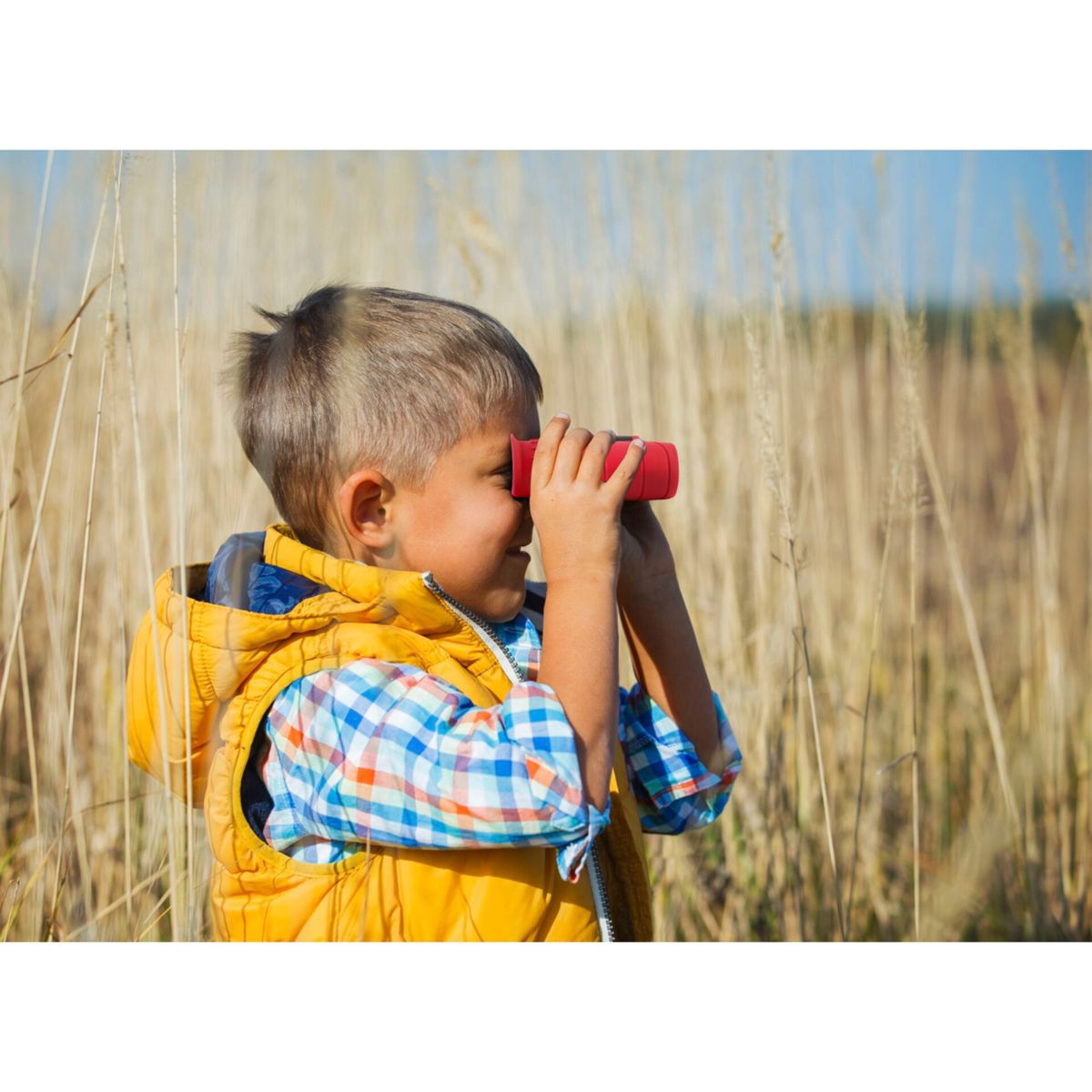 Un ragazzino guarda attraverso un binocolo rosso in un campo di erba alta.
